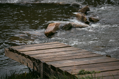 A wooden bridge on the bank of a raging river. Pier by the river. Wooden boards. Rural landscape. Fast mountain river. Water flows through boulders. Stones in the water. Water bubbles. Rapid current.