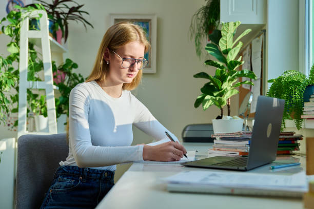 istock Teenage female student sitting at home at desk, writing in notebook using laptop computer 1790095897