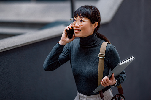 Beautiful Japanese businesswoman, walking outside and going up the stairs. She is smiling while holding a mobile phone and having a phone call. She is looking up and smiling.