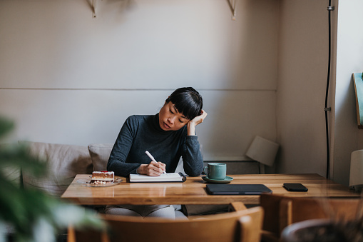 Portrait of a beautiful Japanese woman, sitting in a coffee shop, writing notes in the notebook while having a cup of coffee and a piece of cake. She is looking tired and bored. There is a digital tablet on the table.