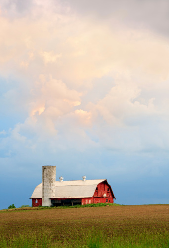 A dramatic sunset sky hangs over a red barn with silo and basketball hoop in the Midwestern United States (Indiana).