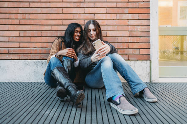 istock Two friends are sitting against a brick wall using a mobile phone together 1790142845