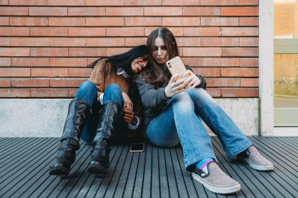 istock Two friends are sitting against a brick wall using a mobile phone together 1790143371