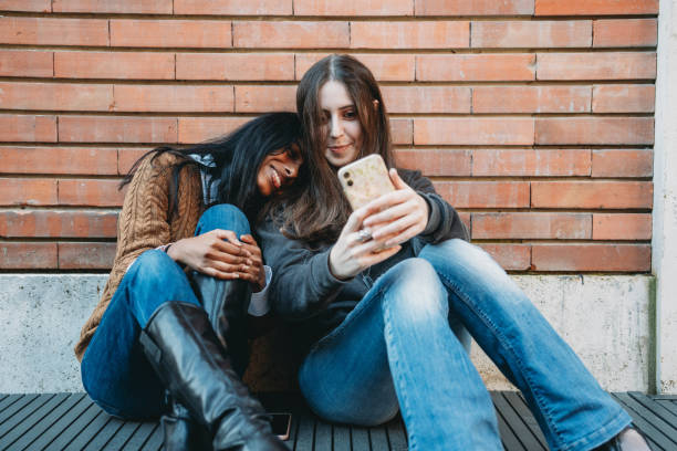 istock Two friends are sitting against a brick wall using a mobile phone together 1790143901