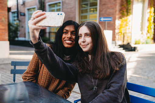 Two friends are taking a selfie together while they are sitting at a table in the city. Sunset time.