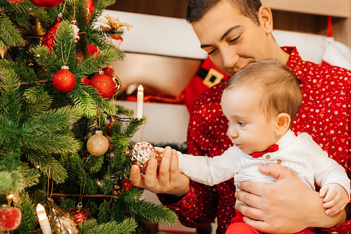 Charming toddler boy in festive clothes near Christmas tree.Christmas picture, tree and lights in the background. Young happy dad and her cute little son are celebrating Christmas or New year.Merry Christmas and Happy New Year.