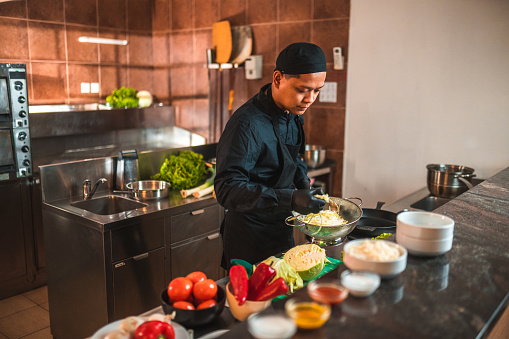 Young asian chef in action, preparing ingredients in the asian kitchen. He is dressed in a black uniform, wearing gloves, and surrounded by a variety of fresh vegetables.