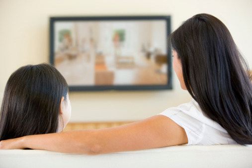 Woman and girl in living room with flat screen television sitting on sofa watching TV