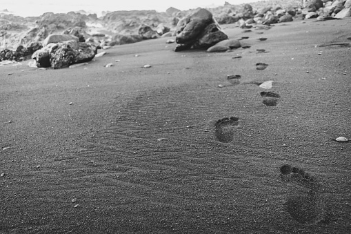 Black and white bare footprints in black, wet sand, walking off into the distance. Volcanic rocks in the background.