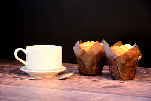 A cup of hot coffee on a saucer and two fresh muffins on a wooden table. Close-up.