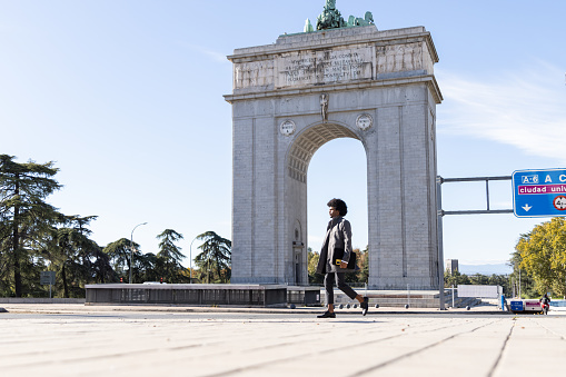 Latin businessman in formal clothing walking through the victory arch in Madrid with his laptop under his arm.