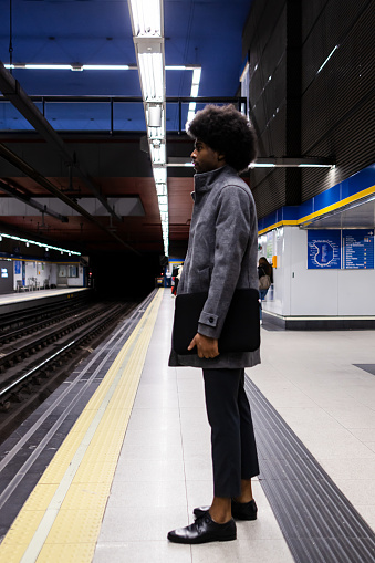 An afro latin businessman is waiting for the subway. He is wearing a gray trench coat and is holding his laptop under his arm. Vertical photo.