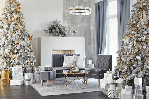 Two large designer snow tree in lounge with lights and balls in white, grey, gold and silver colors. Hall room interior with couch, table, fireplace and decorated luxury Christmas tree with gifts.