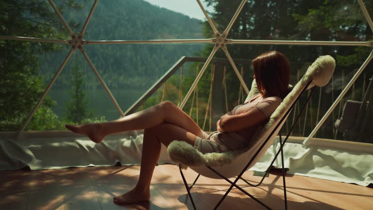 Woman sits near panoramic window overlooking forest and lake