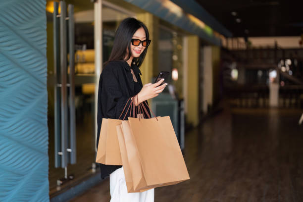 istock Woman using smartphone holding Black Friday shopping bag while standing on the side with the mall background 1790704358