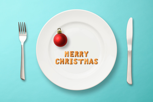 MERRY CHRISTMAS written with biscuits on a white plate, on light blue background.\nWith Knife and Fork