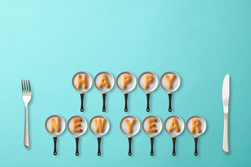 HAPPY NEW YEAR written with biscuits on a frying pan, on light blue background.
With Knife and Fork