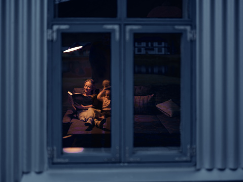Smiling single mother reading a book to her small girl during the night time at home. The view is through window. Photographed in medium format.