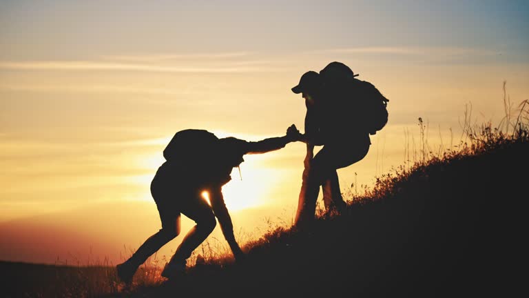 Silhouette two people working in team mountain rock climbing. Helping hand friend to overcome difficulties. Dangerous sport in nature. Business partner. Tourists with backpack and hiking accessories.