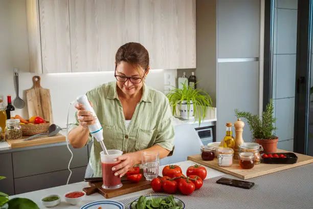 Woman preparing tomato juice iwith immersion blender for a healthy breakfast