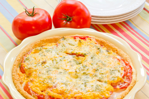 A whole tomato pie topped with cheese, green onions and parsley with tomatoes in the background