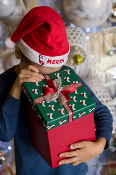 happy little boy wearing santa hat, holding present gift box near christmas tree and looking, peeking inside. merry christmas and happy holidays - christmas child little boys peeking imagens e fotografias de stock