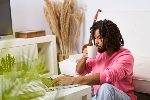 smart young man sitting on floor in living room, working on laptop, drinking coffee