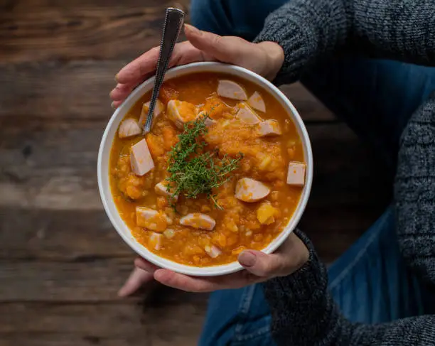 Healthy homemade carrot potato soup or stew with sausage holding by womans hand with warm pullover and jeans sitting crossed legged on wooden floor. Top view