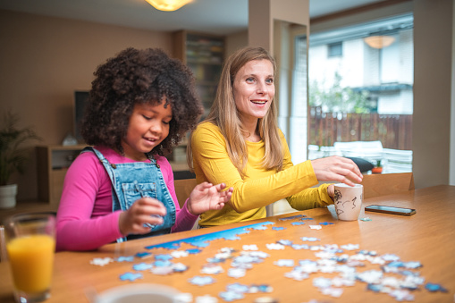A Caucasian mother and her mixed race daughter sit at the dining room table, having fun with puzzles enjoying time together.