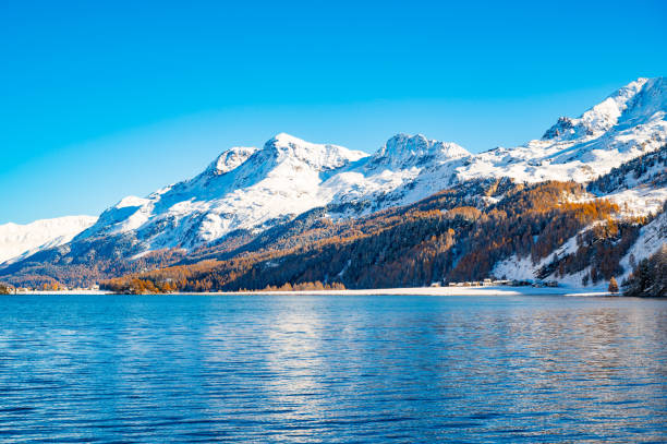 The sun shines brightly over a serene mountain lake, surrounded by autumn-colored forests and snow-tipped peaks under a clear sky.