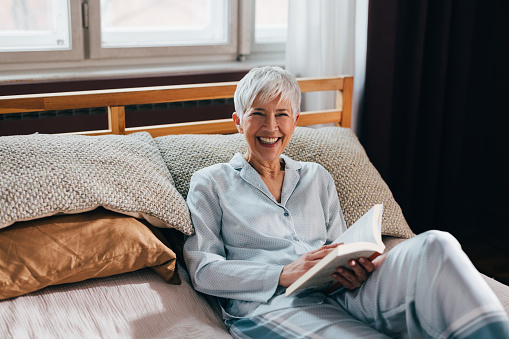 A beautiful senior Caucasian woman lies comfortably on her bed, dressed in cozy pajamas, engrossed in a fascinating book.