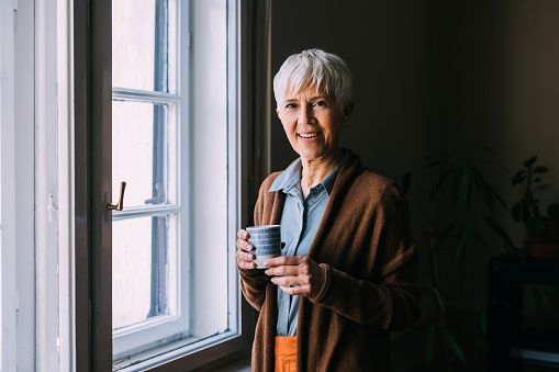 A reflective moment unfolds as a senior Caucasian woman stands by the window, her thoughts carried away by the scenes outside. Holding a cup of hot beverage, she savors the moment, finding comfort in the ritual of sipping.