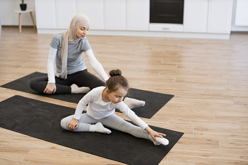 Top view of small female kid on yoga mat stretching arms and legs with slim muslim lady with closed eyes during training. Young parent and daughter in sportswear spend time happily during workout.