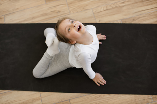Top view of preschool girl in sports wear is engaged in gymnastics at home.Sporty cute female sitting on mat and stretching by touching tips of legs to head in bright spacious room , copy space.
