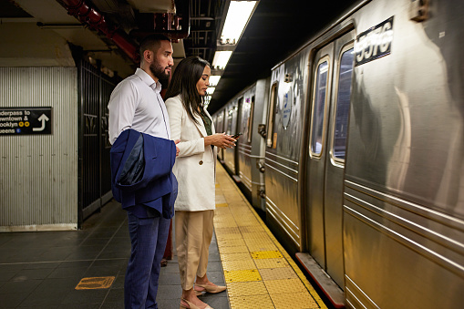 Side view of New Yorkers in summer business attire standing on platform and using smart phone as cars move through station.