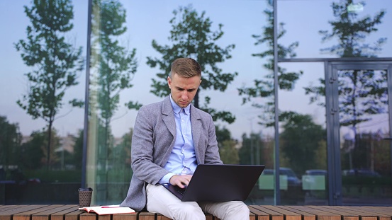 Businessman working outdoors, sitting on the bench, typing on laptop. Copy space. Business, technology concept. Slow motion