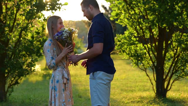 Romantic date in park. Caucasian handsome joyful guy giving flowers to young beautiful woman outdoor. Summer weekend on nature. Love relationships. Girlfriend hugging her boyfriend. Real time.