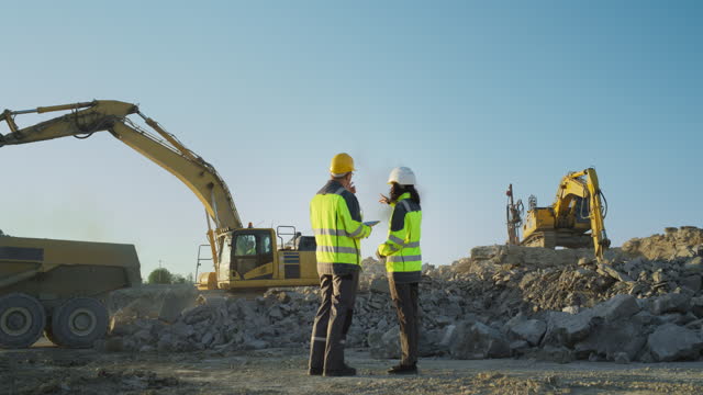 Back View Of Hispanic Female Project Manager And Caucasian Male Civil Engineer Walking and Discussing Real Estate Project On Construction Site. Excavators Digging The Ground For Building Foundation