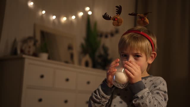 Merry Christmas. Funny child boy drinking milk and having fun. Happy New Year. Portrait of excited little kid wearing pajamas sitting on the bed, making face. Cute baby in a red carnival deer headband