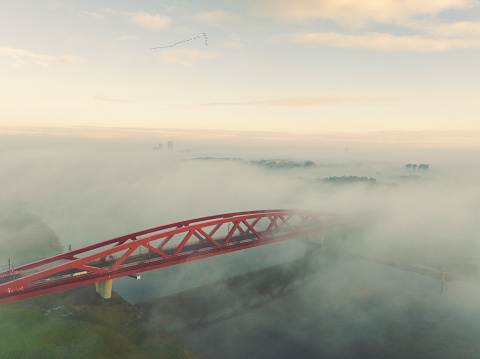 River IJssel covered in a blanket of mist seen from above at the Hanzeboog train bridge during a fall morning near the river IJssel in Overijssel. The treetops are popping out over the thick fog with some of the trees starting to change color during the annual autumn season. The fog is giving the landscape a mystical atmosphere.