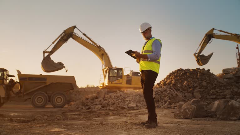 Caucasian Male Real Estate Investor Wearing Protective Goggles And Using Tablet On Construction Site On A Sunny Day. Man Inspecting Building Progress. Excavator Loading Materials Into Industrial Truck