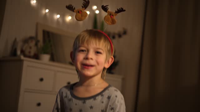 Merry Christmas. Funny child boy laughing and having fun. Happy New Year. Portrait of excited little kid wearing pajamas, jumping on the bed, making face. Dancing baby in a red carnival deer headband