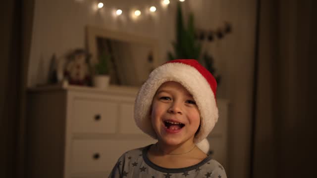 Merry Christmas. Funny child boy laughing while holding Xmas gift box, having fun. Happy New Year. Portrait of excited little kid wearing pajamas, jumping on the bed. Dancing baby in a red Santa hat.