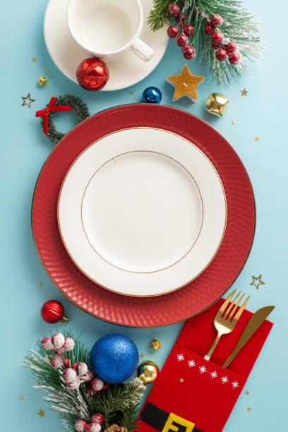 istock Joyful Christmas table arrangement: vertical top-down view featuring plates, whimsical cutlery holder, mug, balls, confetti, frosted fir, holly berries on pastel blueâ space for text or advertisement 1791029866
