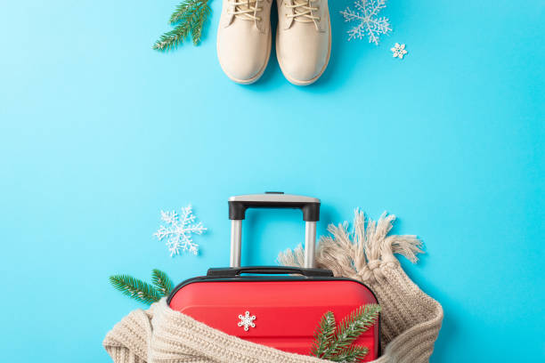 istock Festive getaway notion. A top-view image featuring red suitcase, winter boots, a knitted scarf, fir branches, snowflakes against a bright blue background with space for text or advertising 1791029880