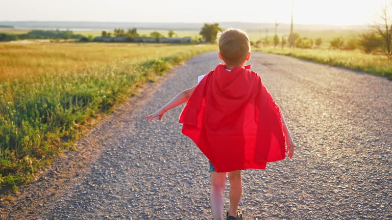 Happy boy child running in superhero costume with red cape on agricultural road in field outside city under sunny summer rays. Boy has spread his arms to sides and feels sense freedom