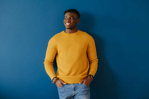 Happy young African man in casual wear holding hands in pockets while standing on blue background