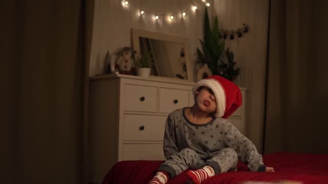 Merry Christmas. Funny child boy laughing while holding Xmas gift box, having fun. Happy New Year. Portrait of excited little kid wearing pajamas, jumping on the bed. Dancing baby in a red Santa hat.