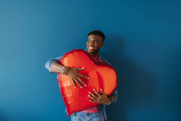 istock Handsome young African man holding heart shaped balloon and smiling while standing on red background 1791039080