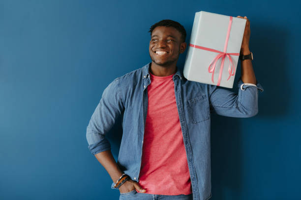 Handsome young African man in casual wear carrying gift box on shoulder and smiling on blue background
